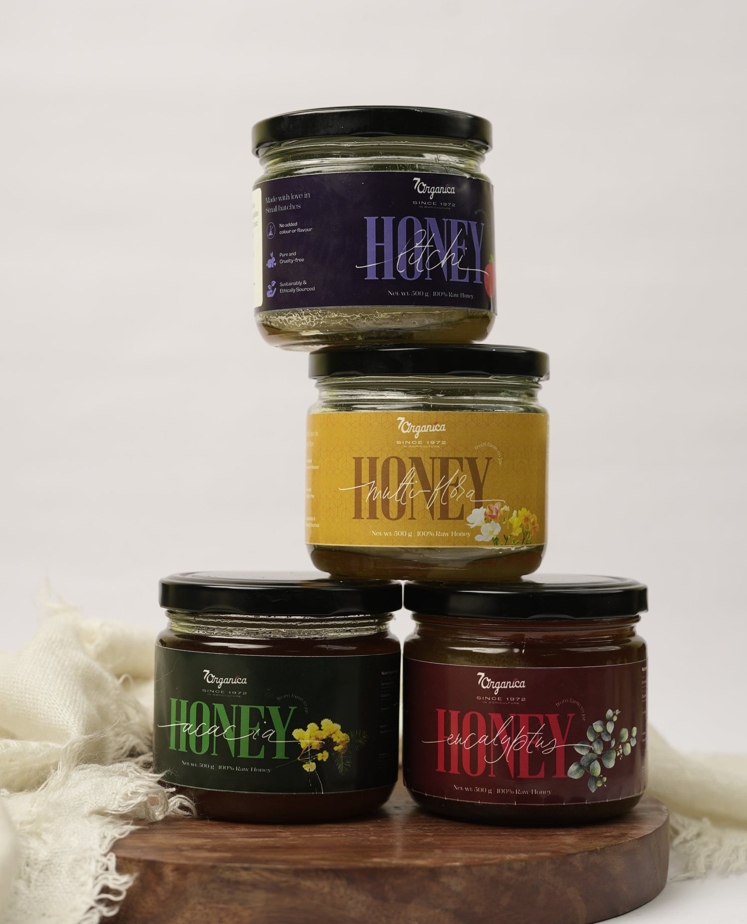 Four jars of honey stacked on a wooden board with white fabric background